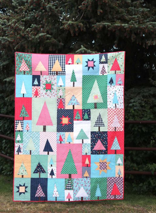 New Patchwork Forest Quilt Pattern: Pine Hollow Version by popular quilting blog, Diary of a Quilter: image of a multi-colored patchwork forest tree quilt displayed outside by a large pine tree.