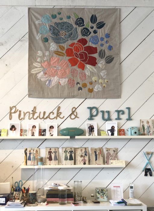 Pintuck & Purl - Modern Fabric and Knitting Shop by popular quilting blog, Diary of a Quilter: image of floral quilt hanging on a wall with some fabric patterns resting on shelves beneath it inside Pintuck and Purl.