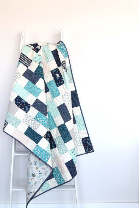 Bricks Baby Quilt Tutorial by popular quilting blog Diary of a Quilter: image of a various shades of blue bricks baby quilt hanging on a white ladder.