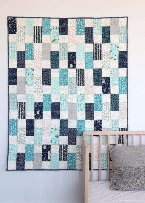 Bricks Baby Quilt Tutorial by popular quilting blog Diary of a Quilter: image of a various shades of blue bricks baby quilt hanging on a wall behind a baby crib.