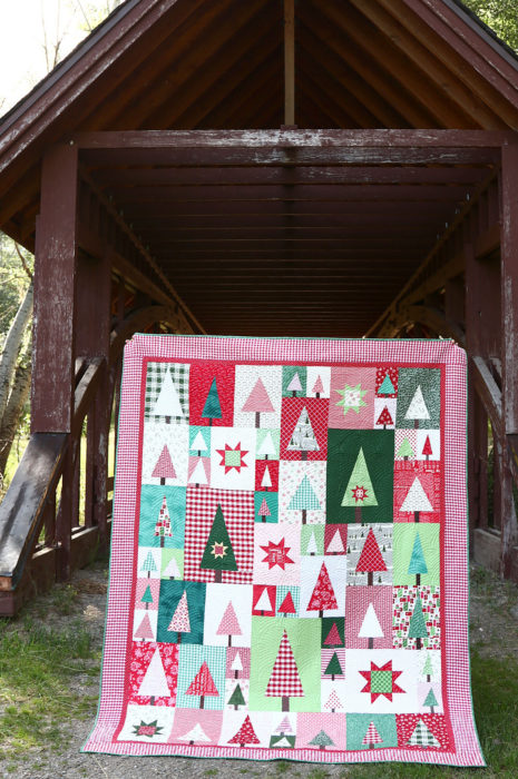 New Patchwork Forest Quilt Pattern: Pine Hollow Version by popular quilting blog, Diary of a Quilter: image of a red, white, and green patchwork forest tree quilt displayed outside in front of a covered bridge.