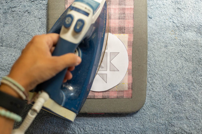 DIY Heirloom Christmas Ornament guest post by Coral + Co by popular Utah quilting blog, Diary of a Quilter: image of a woman ironing an Ohio star template to a square piece of Steam a Seam 2 that is on the backside of a piece of black and red plaid fabric.