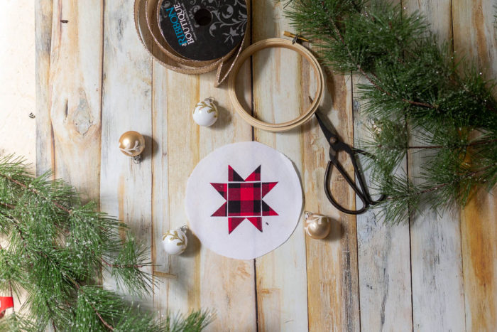 DIY Heirloom Christmas Ornament guest post by Coral + Co by popular Utah quilting blog, Diary of a Quilter: image of a round piece of white felt fabric with a red and black plaid Ohio star on it, embroidery hoop, twine, sewing scissors, and pine tree branches.