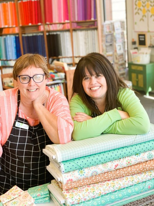 Shop Local - My Girlfriend's Quilt Shoppe + Giveaway by popular Utah quilting blog, Diary of a Quilter: image of two women leaning on top of fabric bolts inside My Girlfriend's Quilt Shoppe.