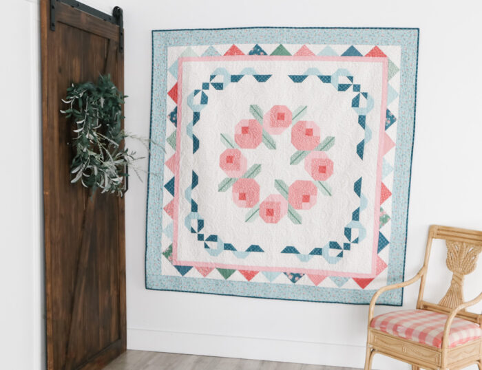 Briarcreek Blooms throw hanging on a wall; light background with floral blocks and tidy pieced frames.